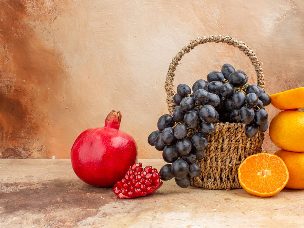 A basket of black grapes beside a pomegranate and fresh oranges